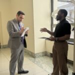 JC stands in a hallway in a congressional office building speaking with a congressional staffer in a grey suit.