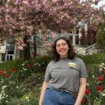 Baylee stands in front of some flowers and a tree on campus